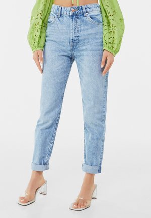 Frau trägt hellblaue, hochgekrempelte Jeans, transparente hochhackige Sandalen und ein grünes, langärmliges, besticktes Oberteil, steht vor weißem Hintergrund.