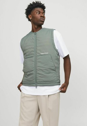 Lichtgroene, gequiltte vest met een rits aan de voorkant, voorzien van een logo op de borst, gecombineerd met een wit T-shirt en beige broeken.
