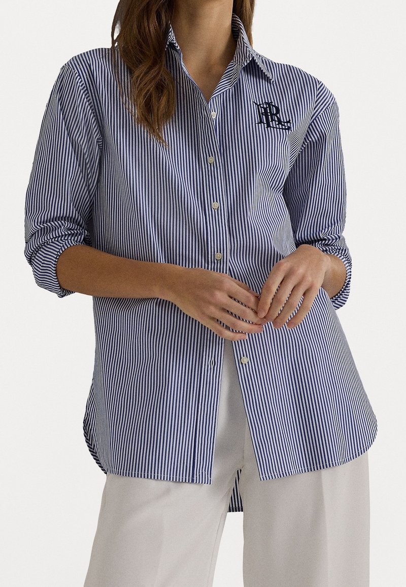 Femme portant une chemise boutonnée rayée verticale bleue et blanche avec manches retroussées sur un pantalon blanc, mains jointes près de la taille.
