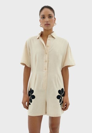 Tuta corta di lino beige con colletto, maniche corte e chiusura frontale con bottoni; presenta ricami neri a forma di fiore sui lati.