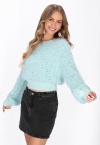 Világoskék szőrös, rövidített pulóver csillogó kiegészítőkkel, fekete farmer mini szoknyával párosítva. A pulóvernek széles ujja van és texturált felülete van.