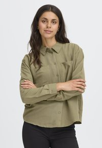 Donna con lunghe capelli scuri, che indossa orecchini a cerchio dorati, una camicia verde oliva con bottoni e pantaloni neri, in piedi con le braccia incrociate su uno sfondo bianco.