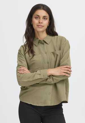Femme aux longs cheveux bruns, portant des boucles d'oreilles en or, une chemise olive à boutons et un pantalon noir, debout avec les bras croisés devant un fond blanc.