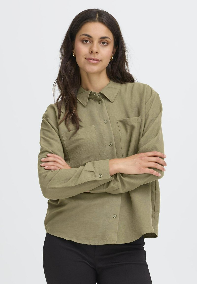 Donna con lunghe capelli scuri, che indossa orecchini a cerchio dorati, una camicia verde oliva con bottoni e pantaloni neri, in piedi con le braccia incrociate su uno sfondo bianco.
