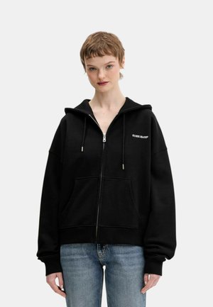 Giovane che indossa una felpa nera con cerniera e il logo "GUESS JEANS" e jeans blu, in piedi contro uno sfondo chiaro uniforme.