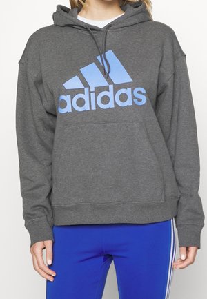 Personne portant un sweat à capuche gris Adidas avec un grand logo bleu et un pantalon de sport bleu roi avec des bandes blanches sur les côtés.