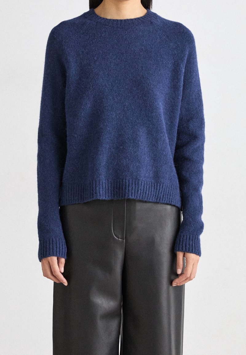 Pull en tricot bleu avec un col et des poignets côtelés, présentant une coupe décontractée et une texture douce, associé à un pantalon noir taille haute.