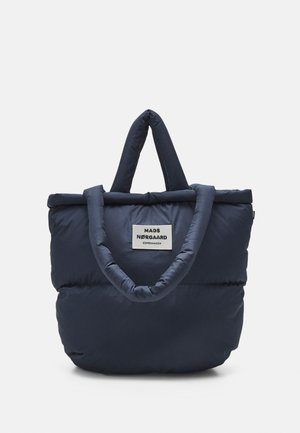 Donkerblauwe gewatteerde draagtas met dikke handvatten en een wit label met de tekst "MADS NØRGAARD COPENHAGEN" op het midden van de voorkant.