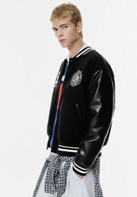 Sort bomberjakke med læderærmer og stribede manchetter, der har et cirkulært logo og knapper, båret over et lag-på-lag-outfit med tern.