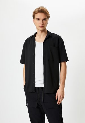 Camisa negra de manga corta en un tejido suave con cuello, botones en la parte delantera y un único bolsillo en el pecho, llevada sobre una camiseta sin mangas blanca.