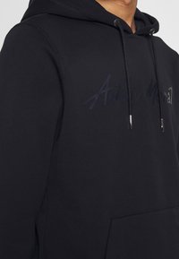 Svart huvtröja i mjukt material, med känguruficka, snörning och en ton-i-ton logotyp på bröstet.