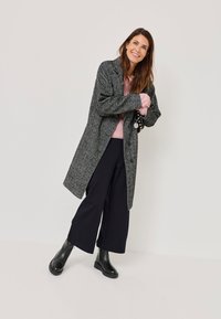 Grau texturierter Mantel mit Kragen, getragen über einem hellrosa Pullover, kombiniert mit weit geschnittenen schwarzen Hosen und schwarzen Stiefeletten.