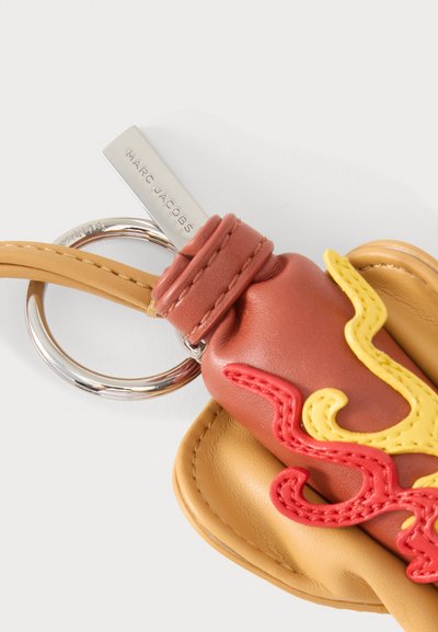 Porte-clés avec des lanières en cuir fauve, une pochette marron avec des flammes brodées jaunes et rouges, et une plaque argentée gravée "MARC JACOBS."