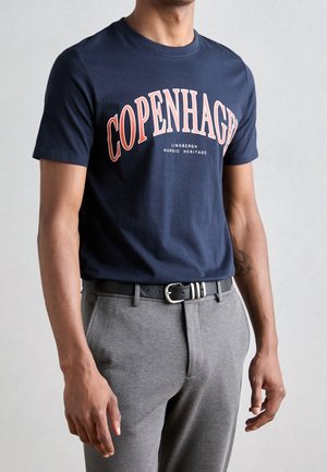 Homme portant un t-shirt bleu marine avec le texte "COPENHAGEN" et un pantalon gris avec une ceinture noire, debout devant un fond clair uni.