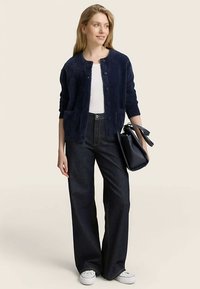 Marineblauer Strickcardigan mit vorderen Knöpfen, getragen über einem weißen Top und dunklen, ausgestellten Jeans, kombiniert mit einer schwarzen Handtasche und weißen Turnschuhen.