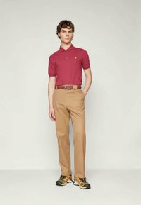 Camisa polo bordô de mangas curtas com um pequeno logótipo, combinada com calças bege e ténis pretos e amarelos. Um cinto castanho destaca o conjunto.