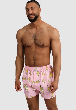 Mann mit Bart und geflochtenem Haar, der rosa Boxershorts trägt, die mit Cartoon-Bananen auf grauem Hintergrund bedruckt sind.