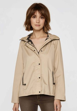 Giovane donna con capelli corti castani che indossa una giacca beige con cappuccio sopra una maglietta floreale e pantaloni scuri, in piedi davanti a uno sfondo uniforme.