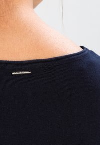 Laivastonsininen neulottu yläosa, jossa on leveä kaula-aukko ja ribbineulos. Takana niskassa pieni metallinen merkki, jossa lukee "CASUAL IDENTITY."