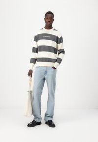 Csíkos pulóver kr�ém és szürke színben, bordás mandzsettákkal, "GUESS JEANS" logóval a mellrészen, világoskék farmerrel és fekete cipővel párosítva, vászontáskával a kezében.