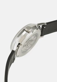 Silberne Edelstahluhr mit poliertem Gehäuse, strukturiertem Aufzugsknopf und schwarzem Lederarmband, mit Glasboden, der feine Details zeigt.