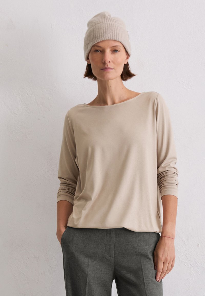 Beige langærmet top med vid halsudskæring og oprullede ærmer, der er parret med grå bukser. Modellen bærer en lys beige strikket hue.