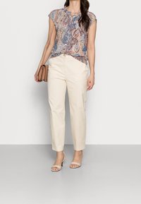 Mönstrad topp i flera färger med kort ärm, kombinerad med krämfärgade byxor och ljusbeiga sandaler. Liten handväska på axeln.