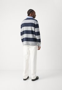 Tengerészkék és szürke csíkos, hosszú ujjú galléros pólóing, kényelmes fazonban. Fehér nadrággal és fekete cipővel párosítva, hátulnézetben.