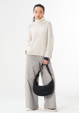 Young woman wearing cream turtleneck sweater and gray check wide-leg pants holds black shoulder bag with strap against white background.