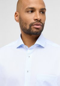 Camicia azzurra chiara con colletto a punta, taschino sul petto e dettagli dei bottoni a contrasto. Tessuto liscio, design aderente.