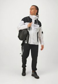 Bela in črna lahka jakna z zadrgo, črne hlače in črni nahrbtnik. Izdelano iz mešanice tekstur in z modernim zunanjim dizajnom.