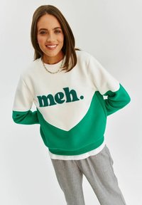 Zeleno-bílý barevně blokovaný svetr s nápisem "meh." v modrozelené barvě. Měkká textura s žebrovanými manžetami a lemem, v kombinaci se světle šedými kalhotami.