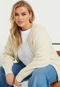 Beige stickad cardigan med ribbade muddar, bärs över en vit T-shirt, parat med blå vida jeans. Tyget har en strukturerad yta.
