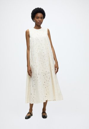 Kvinde iført en ærmeløs, ankel-længde cremefarvet hulmønstret kjole med rund halsudskæring og sorte flade sko, stående mod en ensfarvet baggrund.