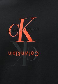 Μαύρο ύφασμα με επικαλυπτόμενα λογότυπα Calvin Klein σε μεγάλα κόκκινα και μαύρα γράμματα, ένα λογότυπο είναι αναποδογυρισμένο.