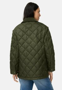 Vrouw in een donkergroene gewatteerde jas met drukknoop manchetten en een brede kraag, gecombineerd met lichtblauwe jeans, gezien vanaf de achterkant.