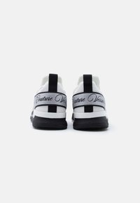 Λευκά αθλητικά παπούτσια με πλέγμα στο πάνω μέρος, μαύρες λεπτομέρειες και αντανακλαστικό ασημί λουράκι στο πίσω μέρος με κείμενο. Μαύρη λαστιχένια σόλα.