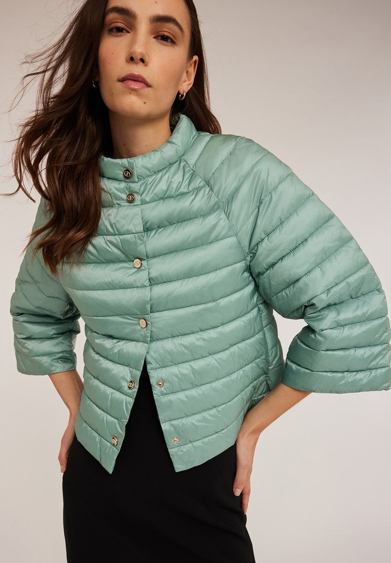 Jasnozielona, puszysta, pikowana kurtka z wysokim kołnierzem i rękawami do trzy czwarte. Posiada guziki z przodu i gładką teksturę.