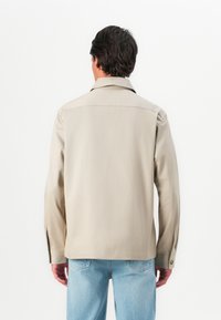 Man wearing beige long-sleeve shirt and light blue jeans, shown from the back against a plain white background.