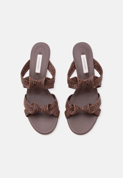 Καφέ πλεκτά δερμάτινα σανδάλια με δύο λουριά και επίπεδες σόλες, με την επωνυμία Casadei, εμφανιζόμενα από πάνω σε λευκό φόντο.