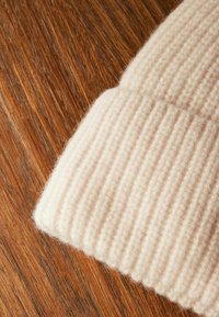 Beige strikket hue med ribbet tekstur. Blødt uldmateriale med en foldet skygge, præsenteret mod en træbaggrund.