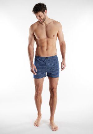 Kaal bovenlijf staande man met kort krullend haar, blootsvoets en gekleed in blauwe korte broek met patroon, kijkt omlaag en glimlacht tegen een effen witte achtergrond.