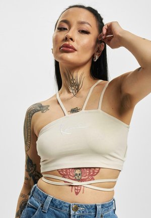 Beige cropped top with thin straps, featuring a fitted design and an embroidered brand name. Notable tattoos visible on model's skin.