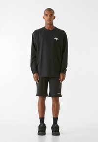 Crna majica dugih rukava sa malim bijelim logotipom na prsima, uparena s crnim šortsevima i crnim tenisicama. Glatka tkanina, opušteni kroj.