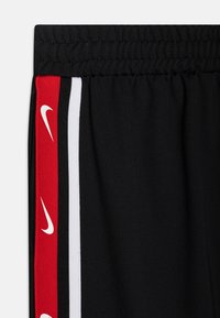 Melnas sporta bikses ar elastīgu jostasvietu, sānu svītrām sarkanā, baltā un melnā krāsā, ar atkārtotām baltām Nike ikonām uz sarkanās svītras.