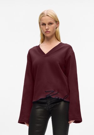Burgunderfarbener V-Ausschnitt-Sweatshirt mit langen Ärmeln, das einen abgerundeten Saum und gesticktes "Karl" in Blau am unteren Ende aufweist. Glatte Stofftextur.