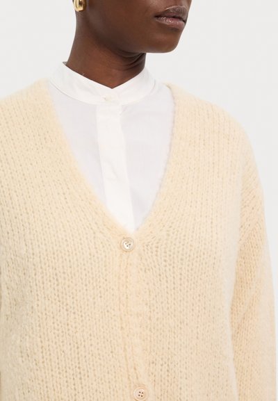 Beige gestrickte Strickjacke mit V-Ausschnitt, ausgestattet mit zwei runden Knöpfen und einem weichen, strukturierten Stoff, der über einem weißen Hemd mit Kragen liegt.