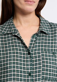 Camicia a quadri verde e beige con colletto, chiusura a bottoni e tasche anteriori con bottoni rotondi. La texture sembra morbida e tessuta.