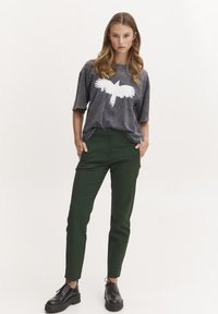Grå oversized t-shirt med et hvidt fugle grafik, parret med tætsiddende grønne bukser og sorte snørebåndssko, der har en glat tekstur.