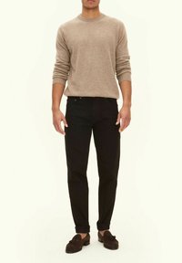 Beiger Strickpullover, schwarze Jeans und dunkle Slipper. Der Pullover hat einen Rundhalsausschnitt und gerippte Bündchen; die Jeans haben eine klassische Passform und dezente Nähte.